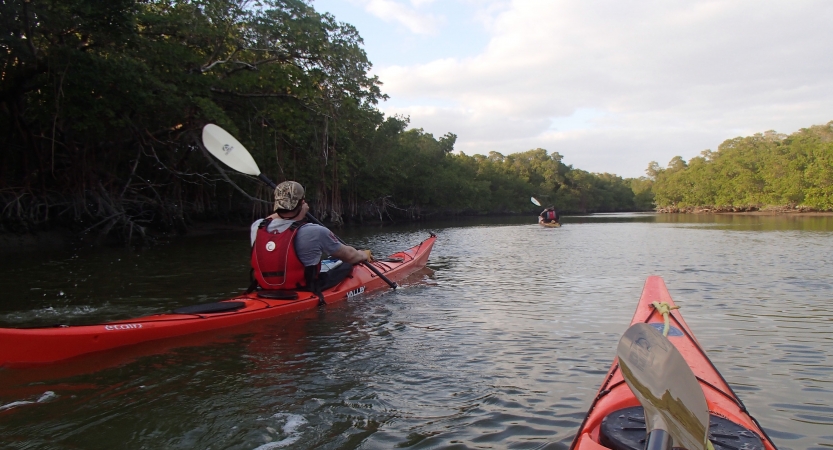 From the view point of a red canoe, a person wearing a life jacket paddles another canoe nearby. 